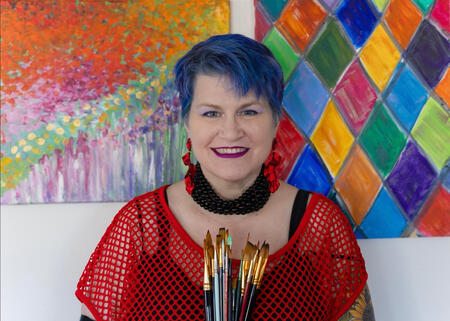 Jodi has short, vibrant blue hair and is wearing bright red mesh top. She smiles at the camera as she stands in front of her bold, colorful artwork. She is holding a bouquet of thin paintbrushes.